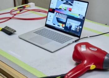 عکس از یک لپ‌تاپ Surface نقره‌ای در حالت باز روی میز تست که صفحه‌نمایش رنگ‌بندی‌های نوار رنگی آزمایشی و نرم‌افزار اندازه‌گیری رنگ و یک پنجره‌ی وب‌کم با حضور افراد را نشان می‌دهد. در سمت چپ میز، یک برس تمیزکردن گرد و غبار مشکی و سیم‌های قرمز و زرد و در سمت راست، یک سشوار صنعتی قرمز با سیم‌های مشکی و زرد قرار گرفته است