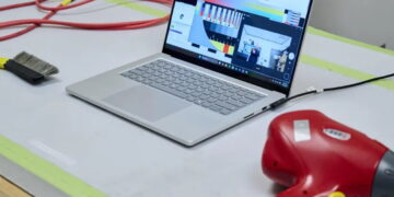 عکس از یک لپتاپ Surface نقرهای در حالت باز روی میز تست که صفحهنمایش رنگبندیهای نوار رنگی آزمایشی و نرمافزار اندازهگیری رنگ و یک پنجرهی وبکم با حضور افراد را نشان میدهد. در سمت چپ میز، یک برس تمیزکردن گرد و غبار مشکی و سیمهای قرمز و زرد و در سمت راست، یک سشوار صنعتی قرمز با سیمهای مشکی و زرد قرار گرفته است