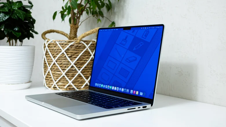 یک MacBook Pro باز روی میز سفید قرار دارد و صفحهنمایش آن رابط کاربری macOS را نشان میدهد.