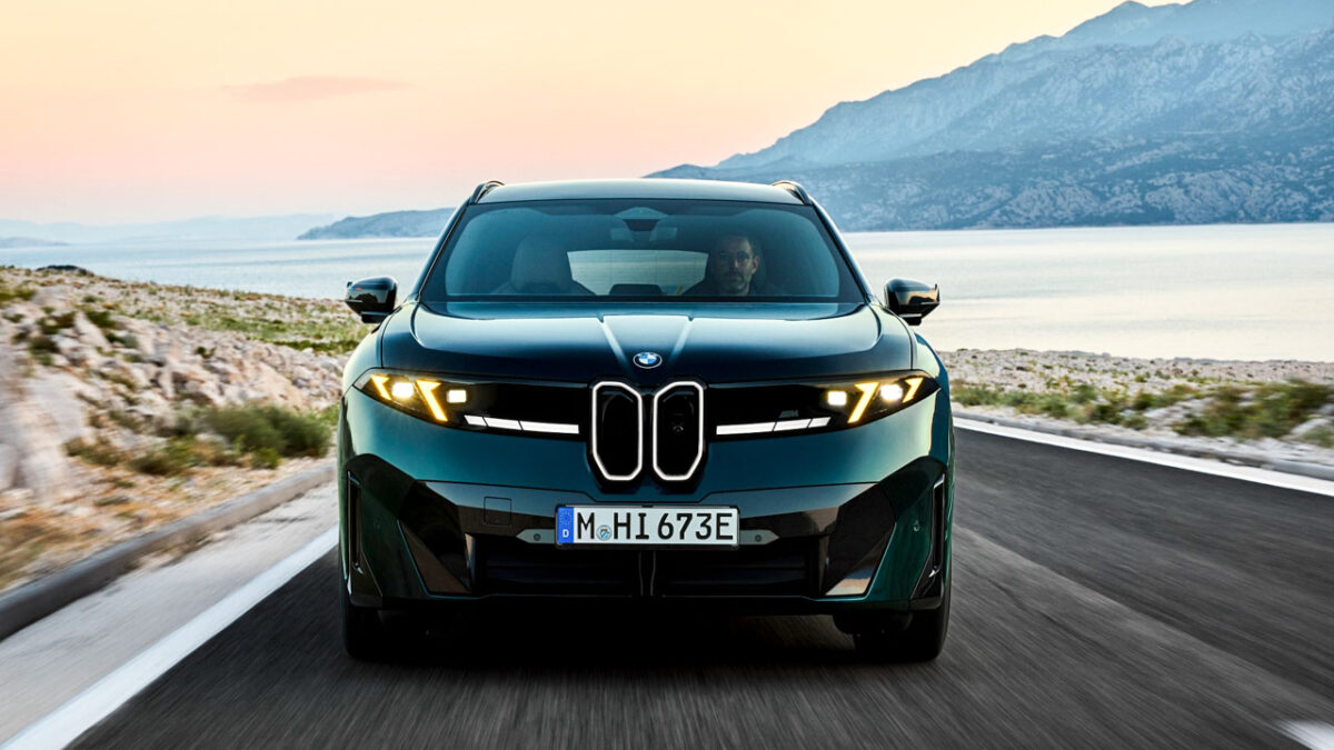 نمای روبهروی BMW iX3 M60 xDrive سبز تیره با چراغهای زرد اختصاصی M، جلوپنجره نورانی و سپر اسپرت در جادهای ساحلی هنگام غروب