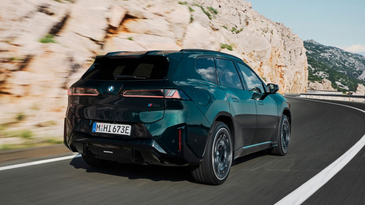 نمای سهرخ عقب خودروی برقی BMW iX3 M60 xDrive سبز تیره با رینگهای اسپرت، چراغهای کشیده عقب و دیفیوزر مشکی در جادهای کوهستانی