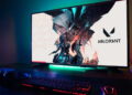 مانیتور گیمینگ با پس‌زمینه‌ی بازی Valorant و نورپردازی RGB در لبه‌ی پایینی، قرارگرفته روی میز در کنار کیبورد و موس مکانیکی با نور پس‌زمینه‌ی قرمز و هدفون گیمینگ