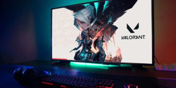 مانیتور گیمینگ با پسزمینهی بازی Valorant و نورپردازی RGB در لبهی پایینی، قرارگرفته روی میز در کنار کیبورد و موس مکانیکی با نور پسزمینهی قرمز و هدفون گیمینگ