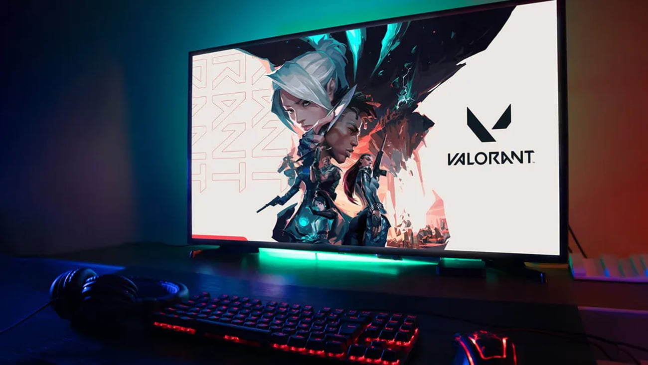 مانیتور گیمینگ با پسزمینهی بازی Valorant و نورپردازی RGB در لبهی پایینی، قرارگرفته روی میز در کنار کیبورد و موس مکانیکی با نور پسزمینهی قرمز و هدفون گیمینگ