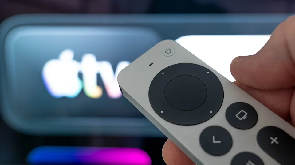 نمای نزدیک از دست فردی که ریموت کنترل نقرهای Apple TV را در دست گرفته و در پسزمینه محو، لوگوی درخشان Apple TV روی نمایشگر دیده میشود.