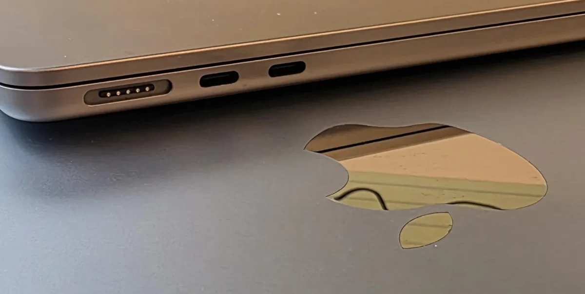 نمای کلوزآپ از لبه کناری یک لپتاپ اپل به رنگ طلایی-قهوهای که پورت MagSafe و دو پورت USB-C روی آن دیده میشود، در کنار سطح مشکی با لوگوی برجسته اپل که انعکاس محیط در آن پیداست.