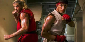 دو مبارز فیلم Street Fighter با لباسهای سنتی قرمز و سفید در حالت رزمی و پشت به یکدیگر در یک سالن تمرینی ایستادهاند.
