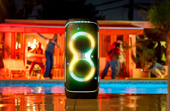 اسپیکر JBL PartyBox مشکی با دو حلقه نورانی رنگین‌کمانی دور بلندگوهایش، ایستاده کنار استخر شنا در شب؛ در پس‌زمینه چند نفر کنار آلاچیق قرمزرنگ در حال رقص‌وشادی هستند و نورهای آبی و نارنجی محیط را روشن کرده است.