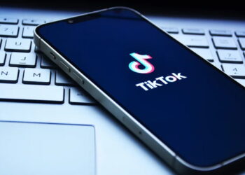 گوشی هوشمندی که روی کیبورد لپ‌تاپ قرار گرفته و لوگوی تیک‌تاک (TikTok) روی صفحه نمایش تیره آن دیده می‌شود.
