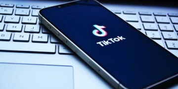 گوشی هوشمندی که روی کیبورد لپتاپ قرار گرفته و لوگوی تیکتاک (TikTok) روی صفحه نمایش تیره آن دیده میشود.