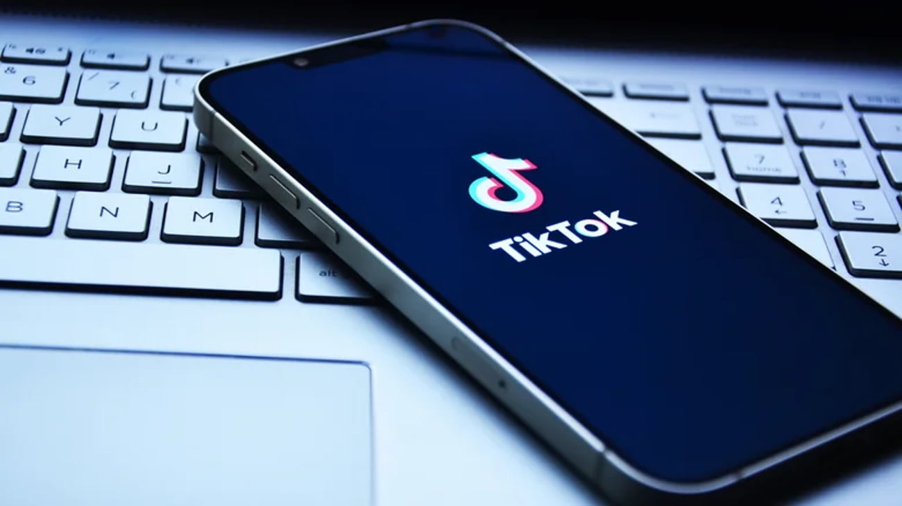 گوشی هوشمندی که روی کیبورد لپتاپ قرار گرفته و لوگوی تیکتاک (TikTok) روی صفحه نمایش تیره آن دیده میشود.