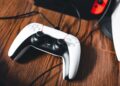 دستهی بیسیم DualSense کنسول PlayStation 5 با کابل شارژ روی سطح چوبی، در کنار نمای محو کنسول و یک Nintendo Switch