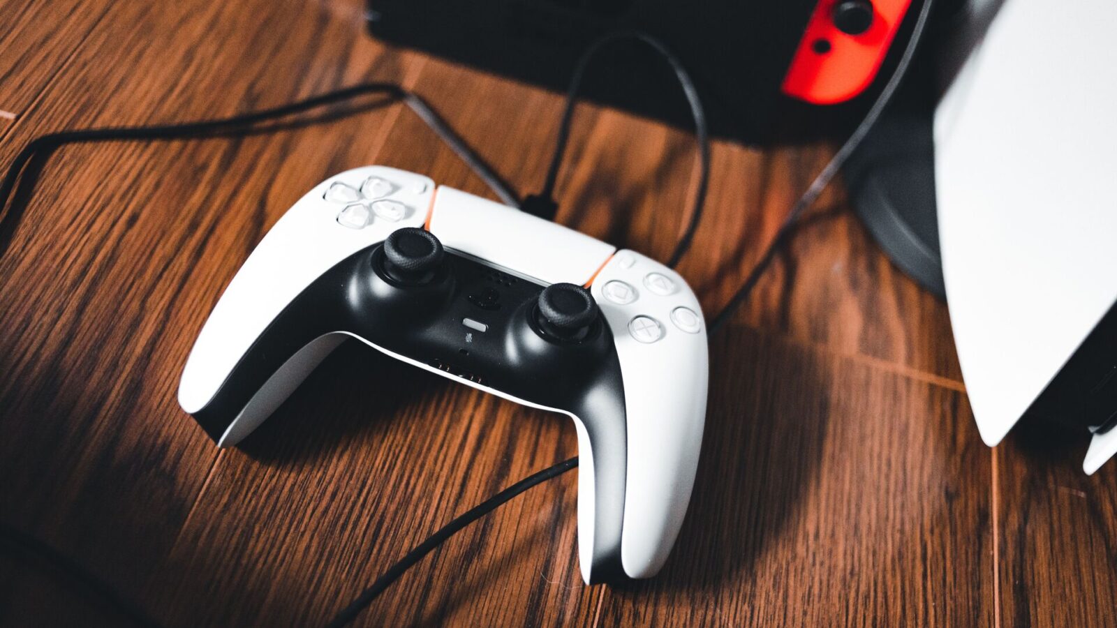 دستهی بیسیم DualSense کنسول PlayStation 5 با کابل شارژ روی سطح چوبی، در کنار نمای محو کنسول و یک Nintendo Switch