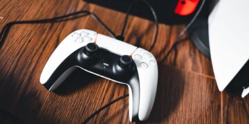 دسته‌ی بی‌سیم DualSense کنسول PlayStation 5 با کابل شارژ روی سطح چوبی، در کنار نمای محو کنسول و یک Nintendo Switch